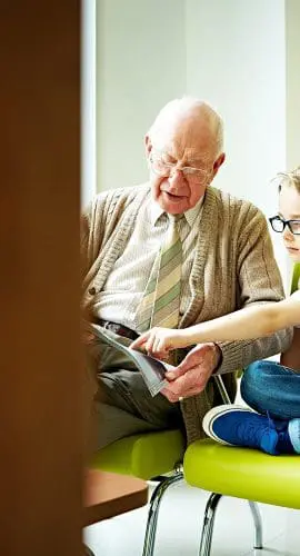 A young boy and an elderly man are sitting together on a green sofa, looking at something the boy is pointing to.