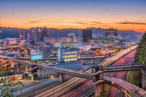 A vibrant cityscape is captured at dusk, featuring illuminated buildings and intersecting highways against a backdrop of rolling hills and a colorful sky.