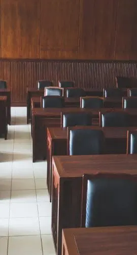 An empty classroom with wooden walls, desks, and black chairs, illuminated by natural light from a window on the side.