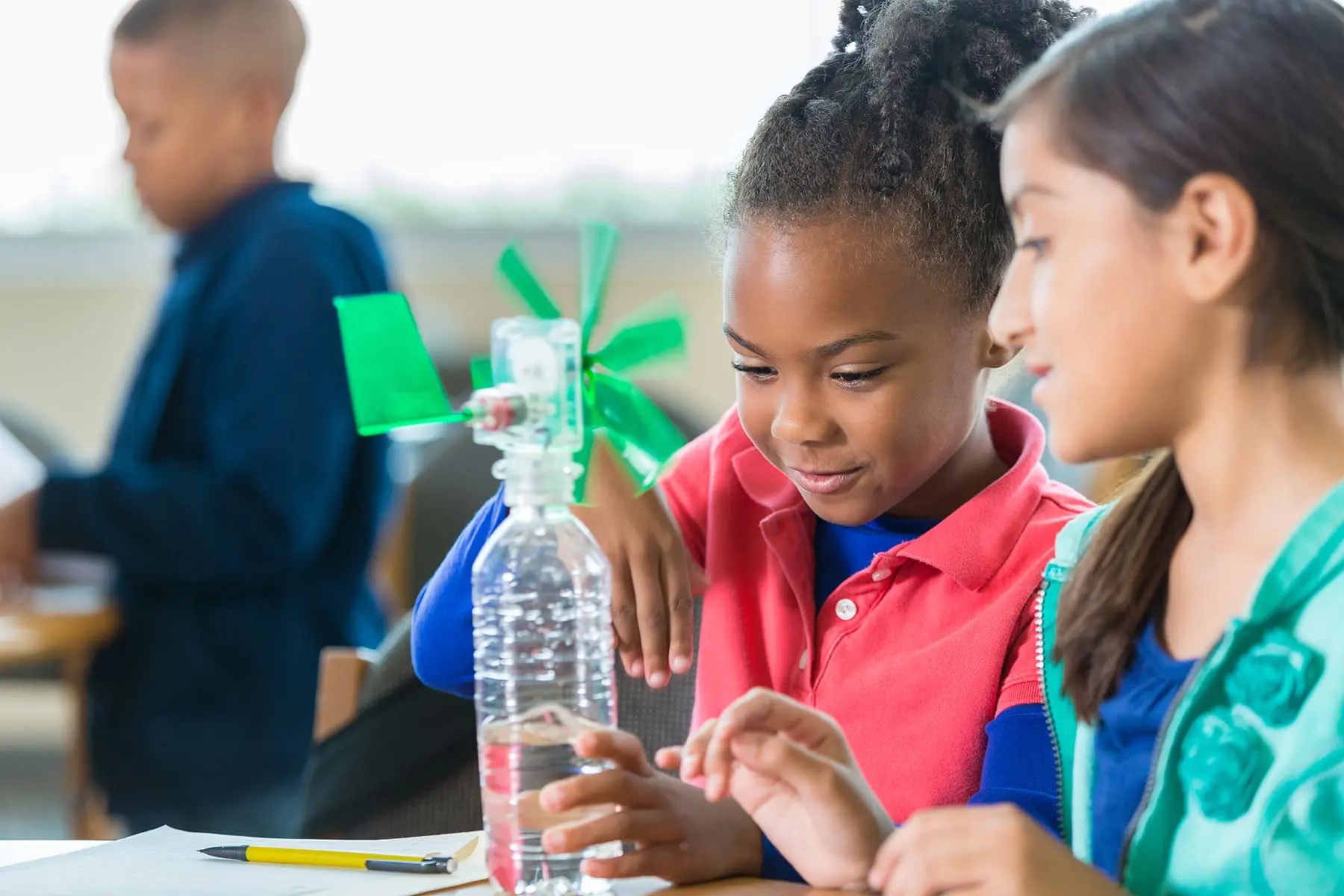 Children are engaged in a classroom activity with a water bottle and paper windmill materials, concentrating on a task at hand.
