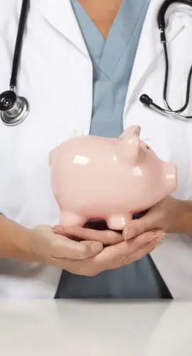 A healthcare professional wearing a lab coat with a stethoscope is holding a piggy bank.