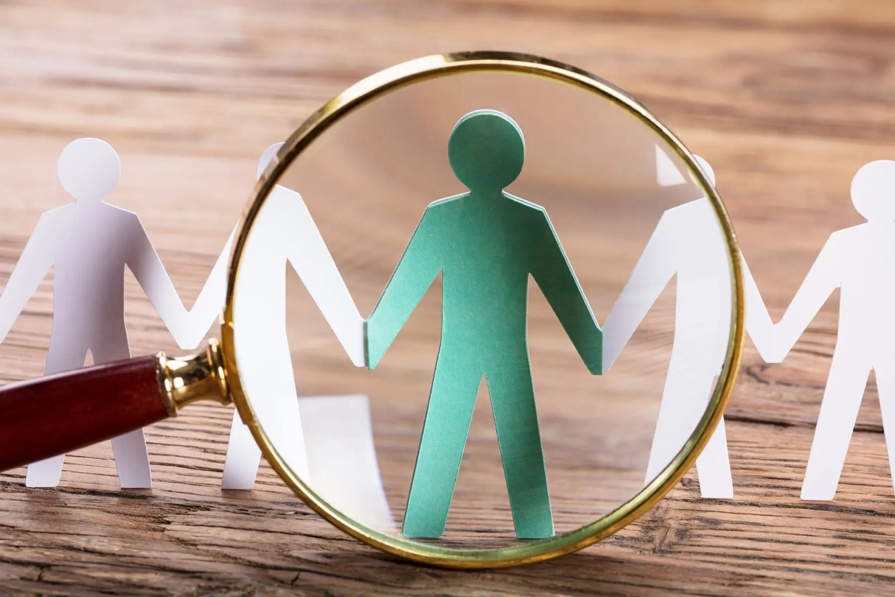 A magnifying glass focuses on a green paper figure among white paper figures, highlighting it.