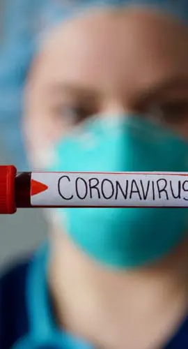 A healthcare worker wearing protective gear holds up a test tube labeled "CORONAVIRUS."