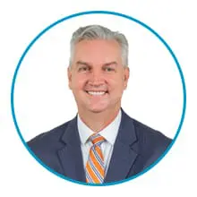 A man dressed in a suit and orange-striped tie, smiling against a white background with a blue circular border.