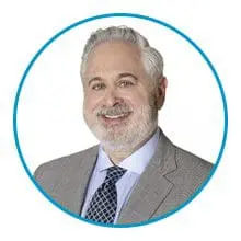 A man with gray hair and a beard is smiling while wearing a suit and tie, framed by a blue circle.