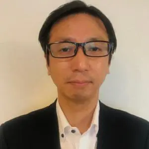 A person with short black hair and glasses is wearing a white shirt and black blazer against a plain background.