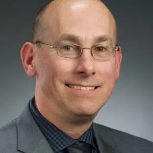 A person is wearing glasses, a suit, and a tie, and is smiling against a plain dark background.