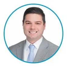 A person is smiling, dressed in a gray suit, white shirt, and light blue tie, inside a blue circular frame on a plain white background.